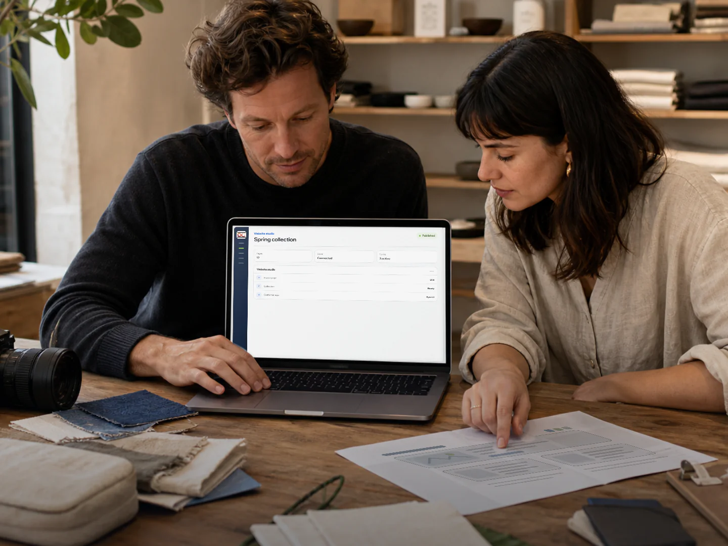 Two people review a website workspace on a laptop beside a printed page layout.