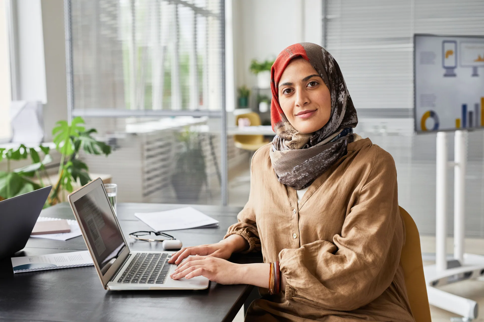 A woman in a headscarf working at a laptop with documents in a bright office.