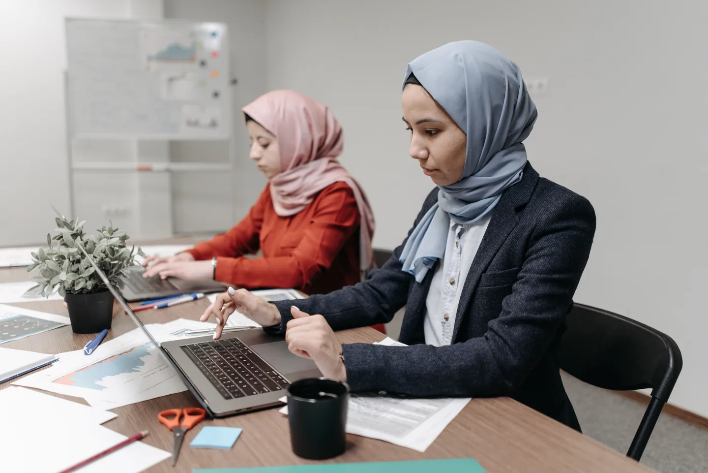 Two colleagues in headscarves working with laptops and documents.