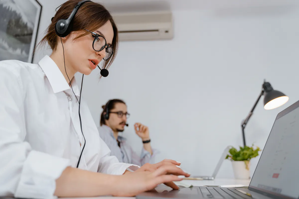 A headset-wearing woman typing on a laptop, with a colleague working behind her.